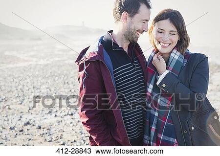 Stock Image - Smiling brunette couple standing on sunny rocky beach. Fotosearch