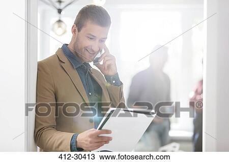 Smiling businessman talking on cell phone and looking down at notebook in office View Large Photo Image Stock Image - Smiling businessman talking on cell phone and looking down at notebook in office. Fotosearch