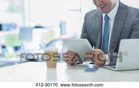 Stock Image - Smiling businessman using digital tablet with coffee in office. Fotosearch