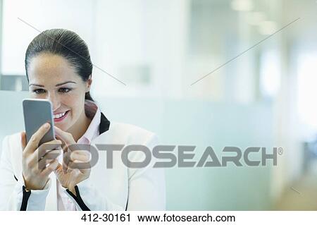 Stock Image - Smiling businesswoman using cell phone in office corridor. Fotosearch
