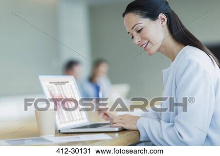 Stock Image - Smiling businesswoman working at laptop with coffee in conference room. Fotosearch
