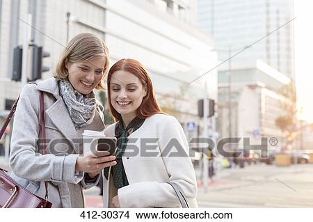 Picture - Smiling businesswomen texting and drinking coffee on city street. Fotosearch