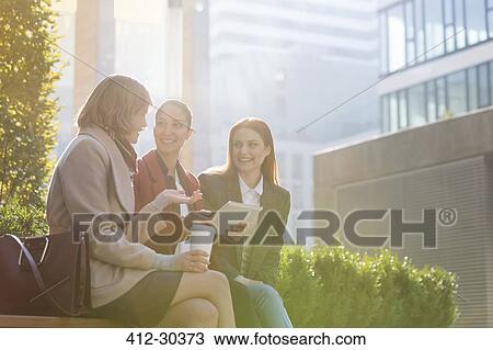 Stock Image - Smiling businesswomen with digital tablet drinking coffee outdoors. Fotosearch