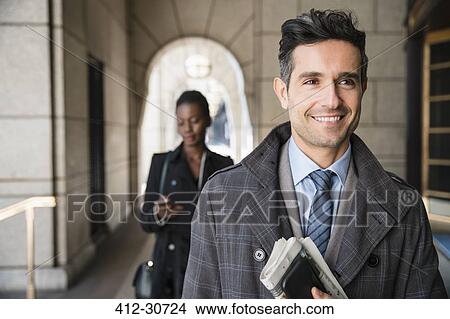 Picture - Smiling corporate businessman carrying newspaper and cell phone in cloister. Fotosearch