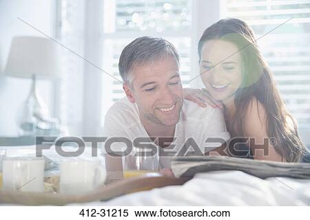 Stock Photography - Smiling couple reading newspaper in sunny bedroom. Fotosearch