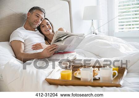 Smiling couple reading newspaper enjoying breakfast in bed View Large Photo Image Picture - Smiling couple reading newspaper enjoying breakfast in bed. Fotosearch