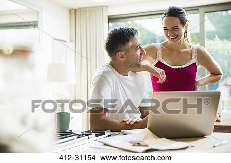 Picture - Smiling couple talking at laptop in morning kitchen. Fotosearch