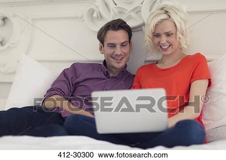Smiling couple using laptop on bed View Large Photo Image Stock Image - Smiling couple using laptop on bed. Fotosearch