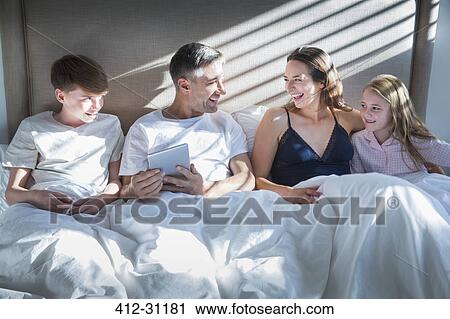 Stock Image - Smiling family laying in bed using digital tablet. Fotosearch