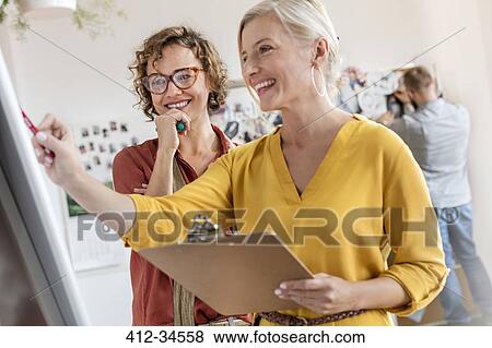 Smiling female design professionals with clipboard meeting at flipchart in office View Large Photo Image Stock Photo - Smiling female design professionals with clipboard meeting at flipchart in office. Fotosearch