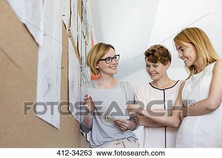 Stock Image - Smiling female designers reviewing proofs at bulletin board in office. Fotosearch