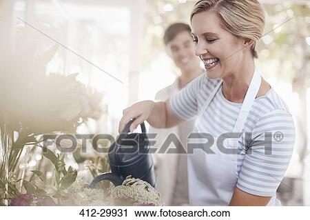 Stock Image - Smiling florist watering flowers in flower shop. Fotosearch