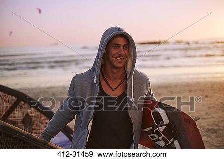 Stock Photo - Smiling man in hoody carrying kiteboard on beach. Fotosearch