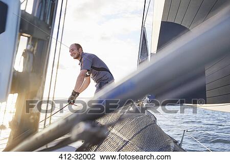 Stock Image - Smiling man sailing holding rigging on sailboat. Fotosearch
