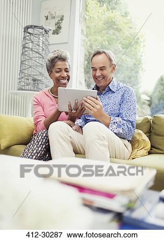 Stock Photo - Smiling mature couple sharing digital tablet on living room sofa. Fotosearch