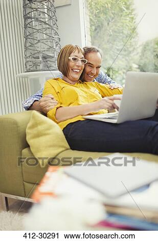 Stock Image - Smiling mature couple sharing laptop on living room sofa. Fotosearch