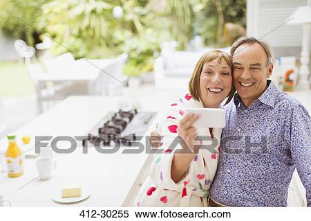 Stock Photography - Smiling mature couple taking selfie in kitchen. Fotosearch