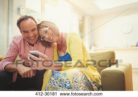 Stock Image - Smiling mature couple texting with cell phone in living room. Fotosearch