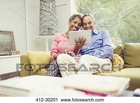 Stock Image - Smiling mature couple using digital tablet on living room sofa. Fotosearch