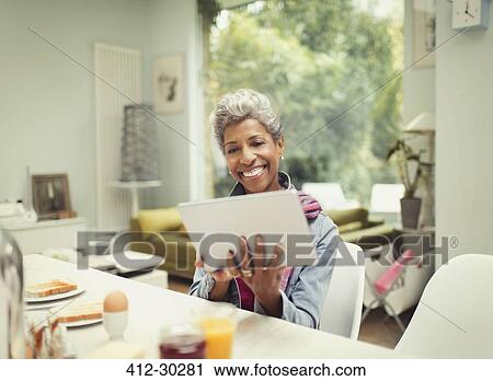 Stock Image - Smiling mature woman using digital tablet at breakfast table. Fotosearch