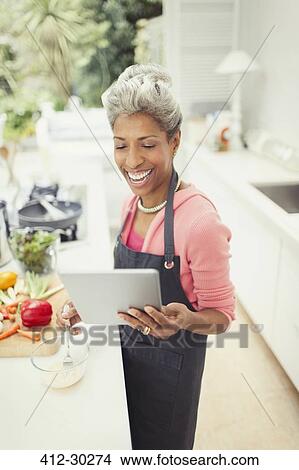 Smiling mature woman with digital tablet cooking in kitchen View Large Photo Image Picture - Smiling mature woman with digital tablet cooking in kitchen. Fotosearch