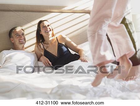 Stock Image - Smiling parents watching children jumping on bed. Fotosearch
