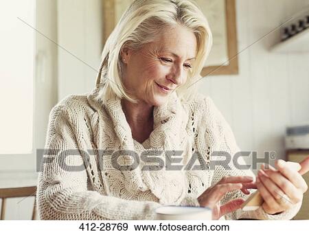 Stock Photo - Smiling senior woman in sweater texting with cell phone in kitchen. Fotosearch