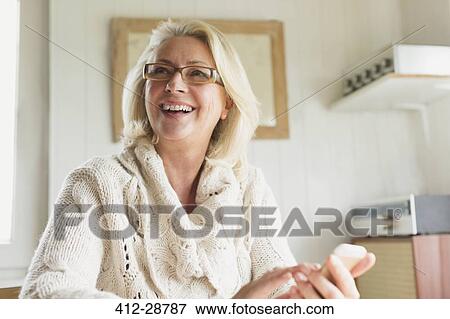 Stock Photo - Smiling senior woman in sweater texting with cell phone in kitchen. Fotosearch