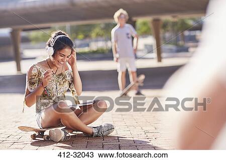 Stock Image - Smiling teenage girl listening to headphones on skateboard in sunny skate park. Fotosearch