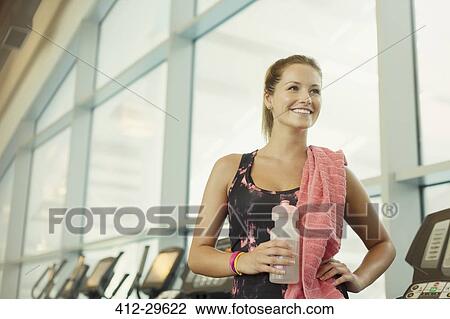 Stock Image - Smiling woman resting and drinking water at gym. Fotosearch