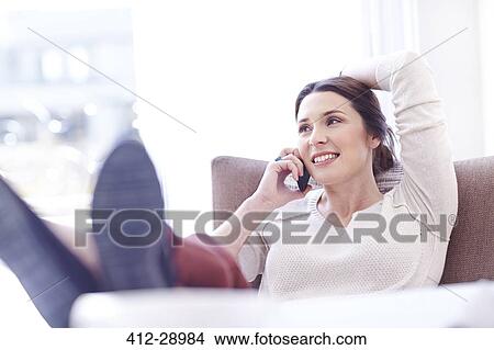 Picture - Smiling woman talking on cell phone with feet up on sofa. Fotosearch