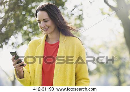 Stock Image - Smiling woman using cell phone in park. Fotosearch
