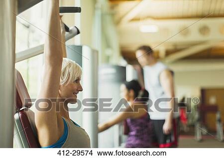 Picture - Smiling woman with arms raised using exercise equipment at gym. Fotosearch