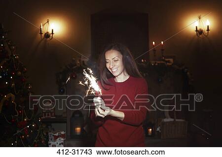 Stock Image - Smiling woman with sparkler firework in dark living room with Christmas tree. Fotosearch