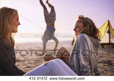Stock Image - Smiling women talking on beach with man doing handstand in background. Fotosearch