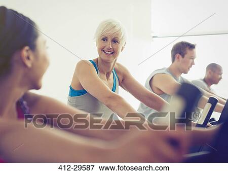 Stock Photo - Smiling women talking on exercise bikes at gym. Fotosearch