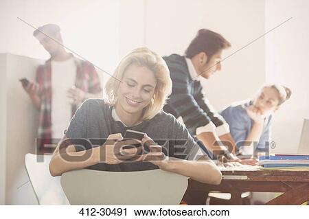 Stock Image - Smiling young businesswoman texting with cell phone in office. Fotosearch