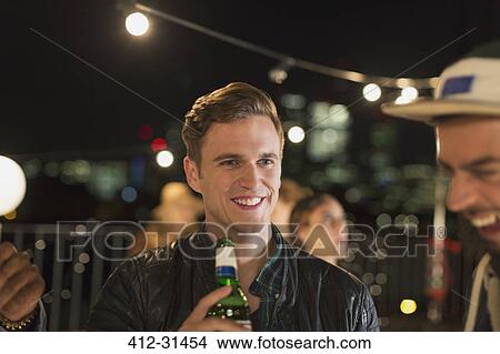 Smiling young man drinking beer at rooftop party View Large Photo Image Picture - Smiling young man drinking beer at rooftop party. Fotosearch