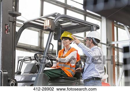 Stock Image - Supervisor directing worker driving forklift in factory. Fotosearch