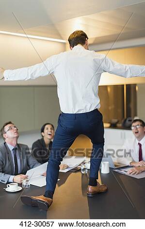 Stock Photo - Surprised colleagues watching exuberant businessman dancing on conference table. Fotosearch