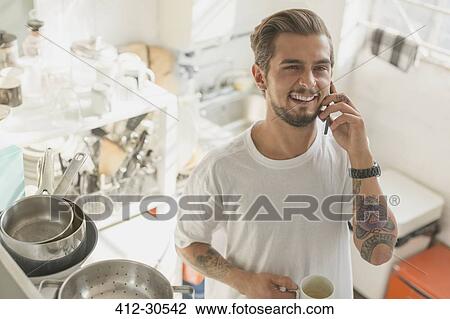 Tattooed young man talking on cell phone and drinking coffee in apartment kitchen View Large Photo Image Stock Image - Tattooed young man talking on cell phone and drinking coffee in apartment kitchen. Fotosearch