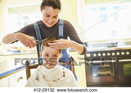 Stock Image - Unhappy boy getting haircut from mother in kitchen. Fotosearch