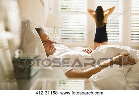 Wife stretching at morning window behind husband sleeping in bed View Large Photo Image Stock Photo - Wife stretching at morning window behind husband sleeping in bed. Fotosearch