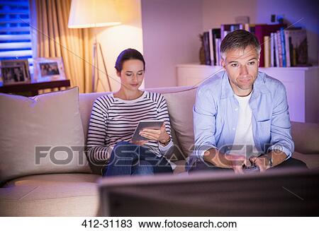 Stock Image - Wife using digital tablet next to husband watching TV in living room. Fotosearch