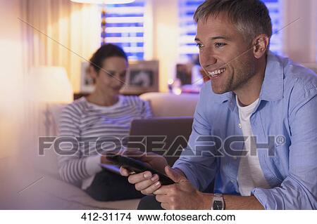 Picture - Wife using laptop and husband watching TV in living room. Fotosearch