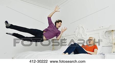 Stock Image - Wife watching playful husband jumping onto bed. Fotosearch