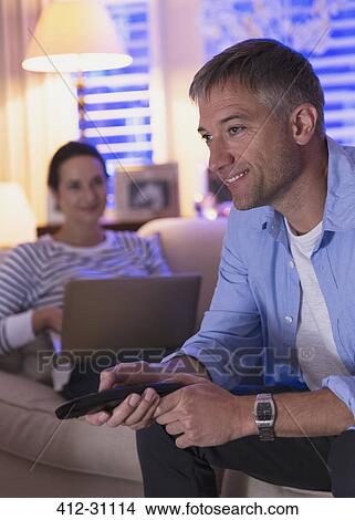 Wife with laptop and husband watching TV in living room View Large Photo Image Picture - Wife with laptop and husband watching TV in living room. Fotosearch