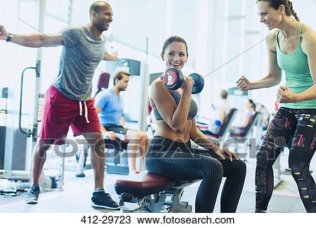 Stock Image - Woman cheering on friend doing dumbbell biceps curls at gym. Fotosearch