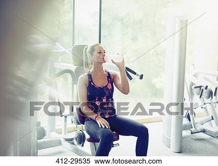 Stock Photography - Woman drinking water at exercise equipment in gym. Fotosearch
