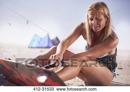Stock Image - Woman fastening kiteboard to feet on sunny beach. Fotosearch